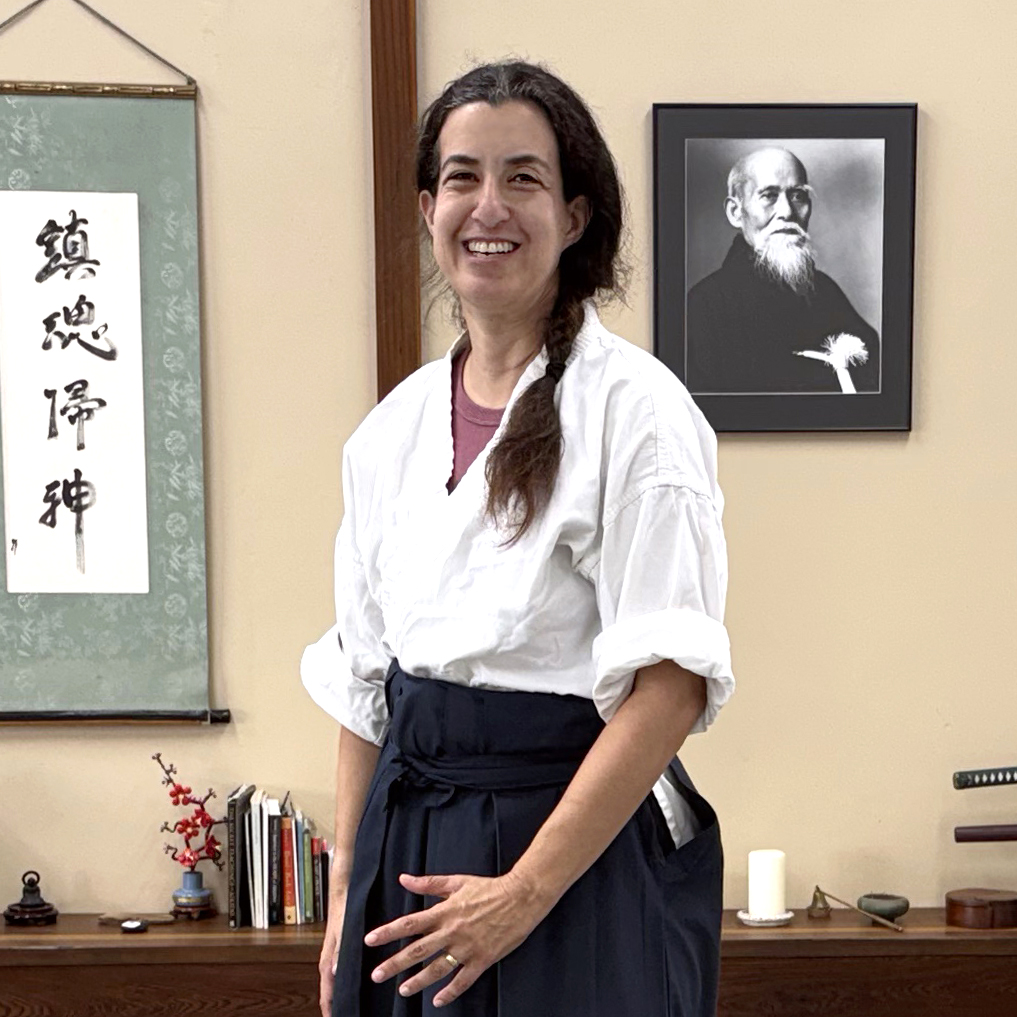 A photo of Adi Elkin, wearing aikido uniform of a white Gi and a black Hakama, standing smiling in front of a photo of Morihei Ueshiba, and Japanese caligraphy. She is excited and smiling.